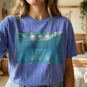 May include: A periwinkle blue t-shirt featuring a graphic of a swimming pool with the text "NO DIVING CAPACITY 0". The shirt is made of a soft material and has short sleeves. The shirt is a comfortable, casual style.
