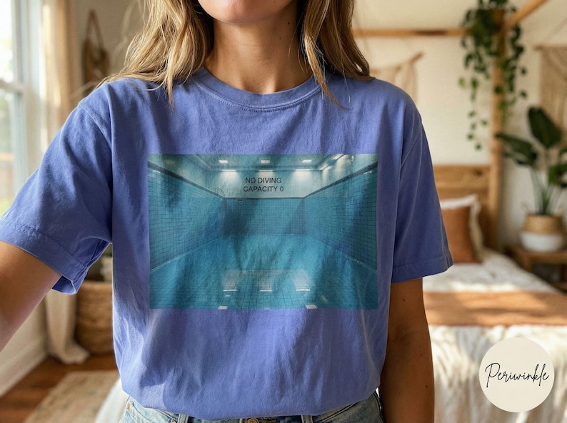 May include: A periwinkle blue t-shirt featuring a graphic of a swimming pool with the text "NO DIVING CAPACITY 0". The shirt is made of a soft material and has short sleeves. The shirt is a comfortable, casual style.