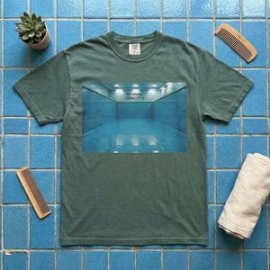 May include: A green t-shirt with a graphic of a blue swimming pool with the text "NO DIVING CAPACITY II". The shirt is on a blue tiled surface, with a small plant, two combs, and a rolled towel.