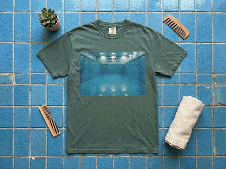 May include: A green t-shirt with a graphic of a blue swimming pool with the text "NO DIVING CAPACITY II". The shirt is on a blue tiled surface, with a small plant, two combs, and a rolled towel.