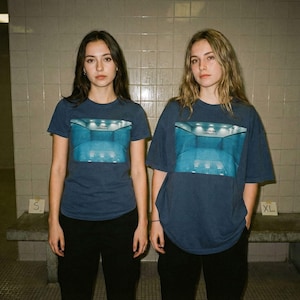 May include: Two people wearing navy blue t-shirts with a blue graphic print. The graphic appears to be a room with a pool. They are wearing black pants. The sizes are S and XL.