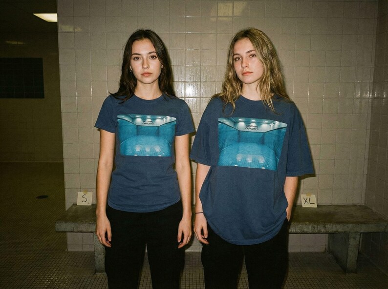 May include: Two people wearing navy blue t-shirts with a blue graphic print. The graphic appears to be a room with a pool. They are wearing black pants. The sizes are S and XL.