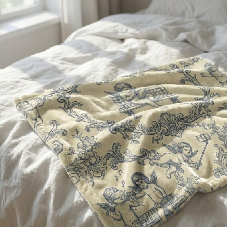May include: A cream-colored throw blanket with a blue and white cherub and floral design. The blanket features cherubs playing musical instruments, using a selfie stick, and relaxing on a bench. The blanket is draped over a white bed.