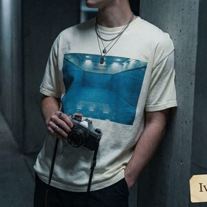 May include: Ivory-colored t-shirt with a blue graphic of a pool and the text "NO DIVING CAPACITY". The person is holding a vintage camera with a black strap. The shirt is a neutral color.