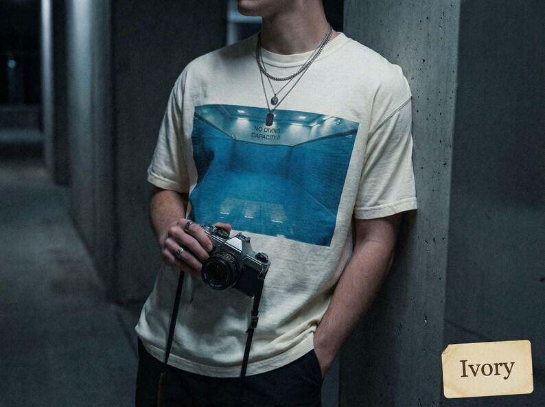 May include: Ivory-colored t-shirt with a blue graphic of a pool and the text "NO DIVING CAPACITY". The person is holding a vintage camera with a black strap. The shirt is a neutral color.