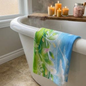 K&ouml;nnte beinhalten: Eine wei&szlig;e Badewanne mit einem blau-gr&uuml;nen Handtuch mit Blumenmuster, das &uuml;ber die Seite drapiert ist. Das Handtuch hat ein Design aus Bl&auml;ttern und Wassertropfen. Kerzen und Badesalze stehen auf einem Holzregal &uuml;ber der Wanne.
