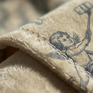 May include: Close-up of a beige fabric with a detailed illustration of a cherub holding a selfie stick. The cherub is depicted with wings and is taking a selfie. The fabric has a soft, velvety texture.