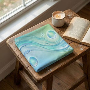 May include: A square, folded fabric with a watercolor design of blue and green water with bubbles. The fabric is resting on a small, rustic wooden stool next to a lit candle and an open book.