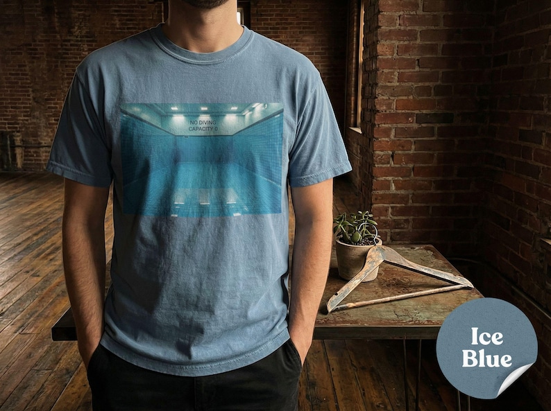 May include: Ice blue t-shirt featuring a graphic of a blue indoor pool with the text "NO DIVING CAPACITY 0". The shirt is a solid color and has a crew neck.