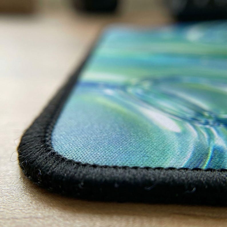 Puede incluir: Primer plano de una alfombrilla de rat&oacute;n rectangular con un dise&ntilde;o abstracto azul y verde. Los bordes est&aacute;n acabados con un borde cosido negro. La alfombrilla tiene una textura suave similar a la tela.
