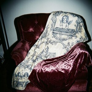 May include: A burgundy velvet armchair draped with a cream-colored throw blanket. The throw features a blue and white illustration of cherubs, musical instruments, and a DJ setup. A matching burgundy velvet blanket is also draped over the chair.
