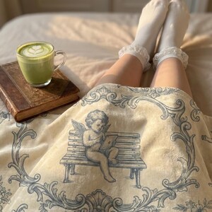 May include: A cozy scene featuring a green matcha latte in a clear glass, a vintage book, and a cream-colored blanket with a blue cherub design. White socks with lace trim are visible. The blanket has a floral and scroll pattern.