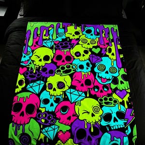 May include: A colorful blanket featuring a repeating pattern of neon green, pink, purple, and blue skulls. The skulls have various designs, including dripping paint, brass knuckles, and diamond shapes, set against a black background.
