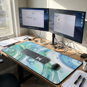 Puede incluir: Un escritorio grande con dos monitores de ordenador, un teclado y un rat&oacute;n. El escritorio tiene una colorida alfombrilla de rat&oacute;n con un dise&ntilde;o de burbujas azules y verdes. Una taza y papeles tambi&eacute;n est&aacute;n en el escritorio.
