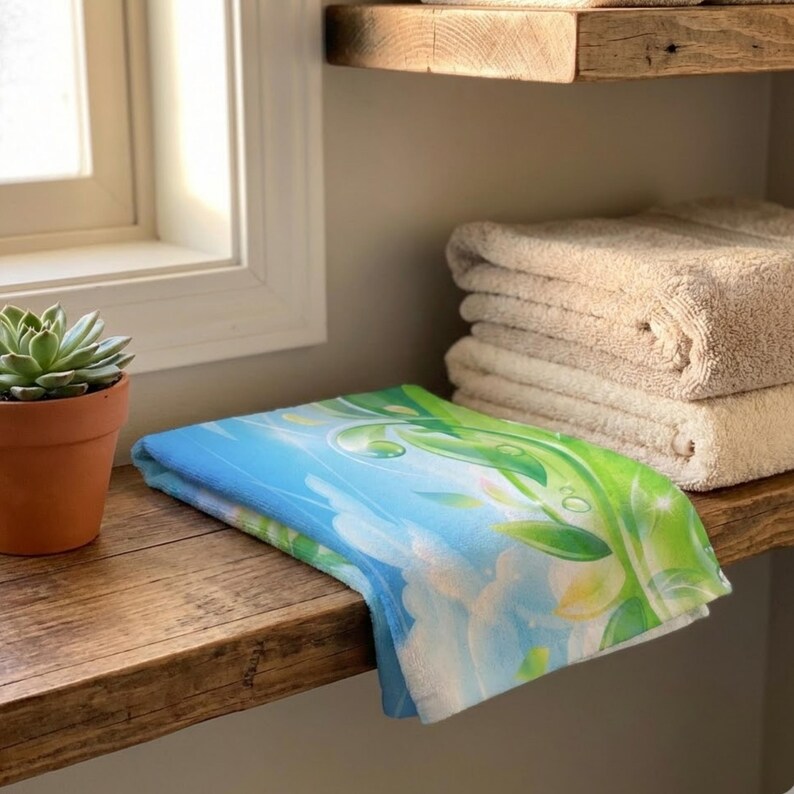 K&ouml;nnte beinhalten: Ein gefaltetes blau-wei&szlig;es Handtuch mit einem Design aus gr&uuml;nen Bl&auml;ttern und Wassertropfen. Das Handtuch liegt auf einem Holzregal neben einer kleinen Topf-Sukkulente und einem Stapel beiger Handt&uuml;cher.