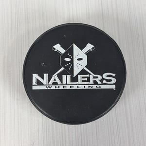 May include: A black circular ice hockey puck featuring a white graphic of a goalie mask with two crossed nails behind it. The text NAILERS is printed in large white capital letters above the word WHEELING in smaller white capital letters.