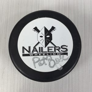 May include: A black ice hockey puck with a white circular center featuring a black graphic of a goalie mask crossed with two sticks. The text reads NAILERS WHEELING with a signature written in silver marker below the logo on the white surface.