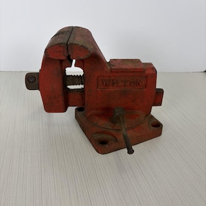 May include: A red metal Wilton bench vise featuring a heavy-duty cast iron body, a threaded spindle, and a swivel base with mounting holes. The tool displays signs of wear on its painted surface, with the brand name Wilton embossed on the side.