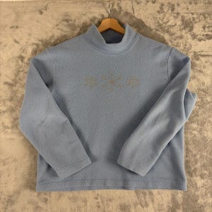 May include: A light blue fleece pullover with a mock neck collar and long sleeves. The center of the chest features an embroidered design of three light-colored snowflakes arranged horizontally across the front of the garment.