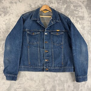 May include: A medium-wash blue denim jacket featuring a pointed collar, long sleeves, and a front button closure. The garment includes two chest pockets with buttoned flaps, vertical seam detailing, and a yellow brand label on the inner neck.