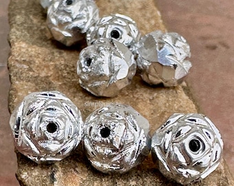 Könnte beinhalten: Nahaufnahme von mehreren silberfarbenen, facettierten Perlen. Jede Perle hat ein Blumendesign mit einem dunklen Mittelloch. Die Perlen sind auf einer rauen, braunen Oberfläche angeordnet. Die Perlen haben eine metallische, reflektierende Qualität.