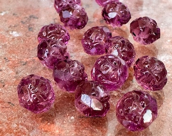 Könnte beinhalten: Eine Sammlung von violetten Amethystperlen mit einem geschnitzten Blumenmuster. Die Perlen sind facettiert und haben eine durchscheinende Optik.