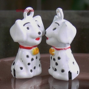 May include: Two white ceramic Dalmatian dog figurines with black spots, red collars, and yellow bells. The figurines are facing each other and have a simple pie logo on the top.