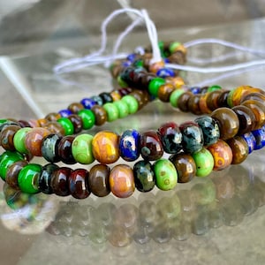 Peut inclure: Un collier de perles en verre multicolores présentant une variété de teintes, notamment vert citron, brun foncé, bleu roi et ambre. Les perles sont rondes et lisses, enfilées sur un fin cordon blanc, créant un ensemble vibrant et brillant.