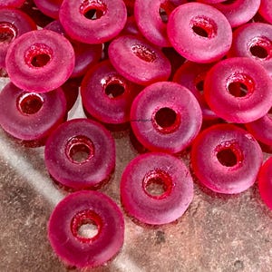 May include: A collection of translucent, ring-shaped beads in a vibrant magenta hue. Each bead has a central hole, and the light passes through them, creating a glowing effect. These beads are likely for jewelry making or crafting.