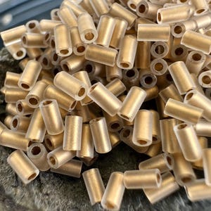 Peut inclure: Une collection de petites perles tubulaires cylindriques avec une finition givrée et semi-translucide dans un ton doré mat. Chaque perle présente un centre creux et une surface extérieure lisse et mate, disposées en un tas dense.