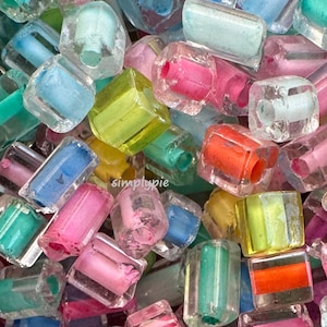 May include: A close-up of a pile of colorful, transparent, rectangular beads. The beads are filled with various colors, including pink, blue, green, yellow, and orange. The beads are scattered, creating a vibrant and playful display.