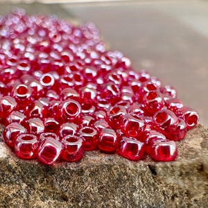 May include: A pile of small, shiny, red beads with a metallic sheen. The beads are scattered on a rough, textured surface. The beads have a hole running through the center, and are likely used for jewelry making or crafts.