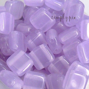 May include: A collection of translucent, light purple, square-shaped beads. The beads have rounded edges and appear to be made of a smooth, polished material. The image is well-lit, highlighting the beads' color and shape.
