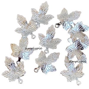 May include: Silver-colored maple leaf charms with detailed veining and a small loop for attaching to jewelry or crafts. The charms are scattered on a white background. The text "simplypie" is visible on some of the charms.