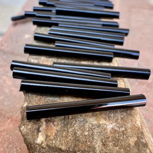 May include: A collection of glossy black glass straws, arranged on a rough, textured surface. The straws are cylindrical and reflect light, creating highlights. The image is a close-up, showcasing the smooth, dark finish of the straws.
