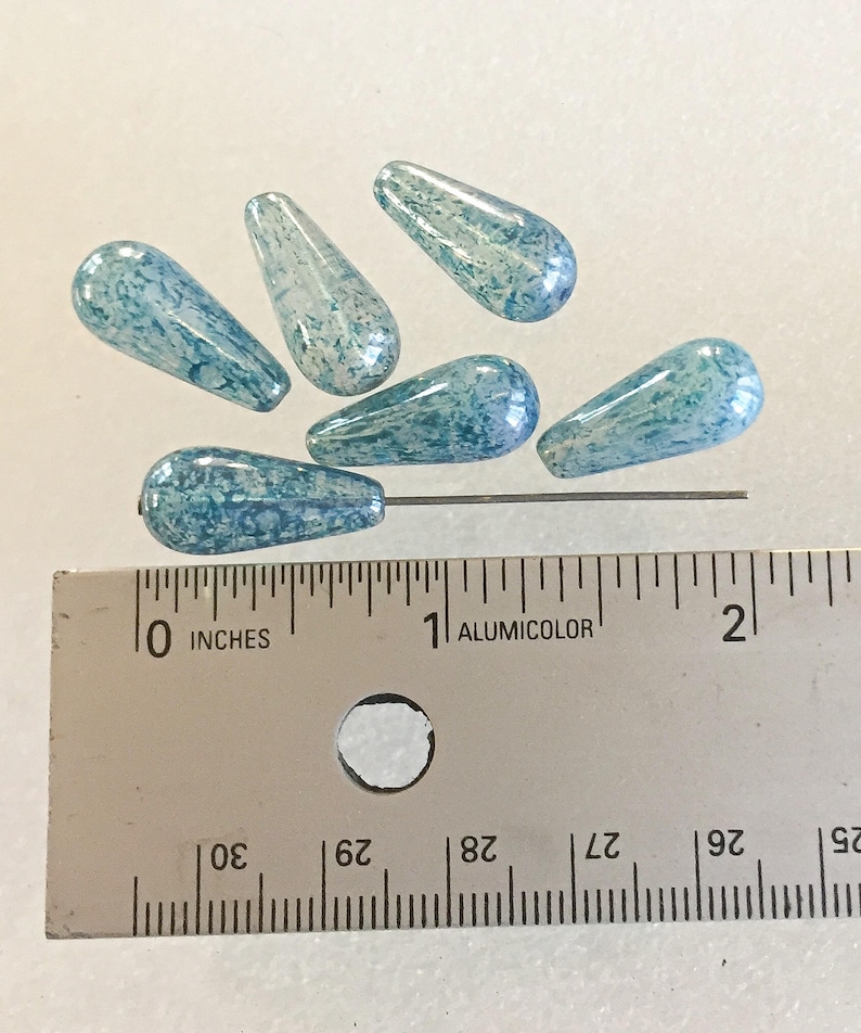 K&ouml;nnte beinhalten: Sechs blau-wei&szlig; tropfenf&ouml;rmige Glasperlen. Die Perlen sind etwa 2,5 cm lang. Ein Metallstift ist durch eine Perle gesteckt. Ein Lineal mit Zollma&szlig;en befindet sich im Vordergrund.