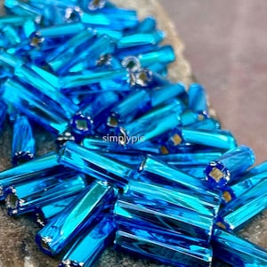 May include: Close-up of a pile of blue glass beads. The beads are cylindrical with a faceted surface, reflecting light. The beads have a metallic silver lining at each end. The word "simplypie" is visible on the surface.