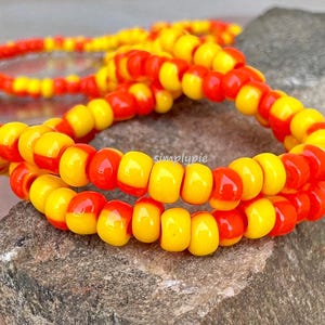 May include: Close-up of two strands of beaded jewelry. The beads alternate between yellow and red, creating a vibrant color contrast. The beads are strung together, forming a necklace or bracelet. The jewelry is displayed on a textured, gray surface.