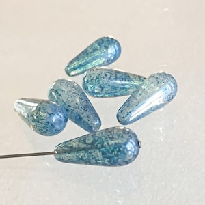 K&ouml;nnte beinhalten: Eine Ansammlung von tropfenf&ouml;rmigen Glasperlen in Blau mit einem gesprenkelten Muster. Die Perlen weisen eine Mischung aus hellen und dunklen Blaut&ouml;nen mit gl&auml;nzender Oberfl&auml;che auf. Eine Perle ist an einer dunklen Metallnadel befestigt.