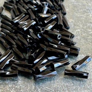 May include: A collection of small, cylindrical black glass beads with a glossy finish. Each bead features a central hole running through its length, and the surface reflects light, highlighting the smooth, dark, and uniform appearance of the pieces.