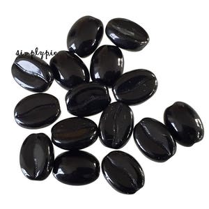 May include: A collection of glossy, black, oval-shaped glass beads, resembling coffee beans, scattered against a white background. The beads have a smooth, reflective surface, and some feature a subtle groove detail. The word "simplypie" is visible in the image.