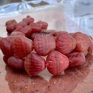 May include: A collection of heart-shaped beads in shades of red and pink. The beads have a textured surface with etched lines. The word "simplypie" is visible in the upper right corner. The beads are strung together, suggesting they are for jewelry making.
