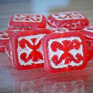 May include: Red glass beads with a white geometric design. The beads are square shaped and have a glossy finish.