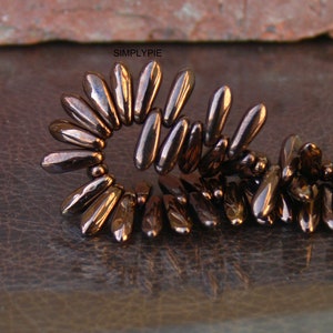 May include: A close-up of a strand of dark brown glass beads. The beads are shaped like long, narrow teardrops and are arranged in a spiral pattern. The beads are shiny and reflective, and the light catches the edges of the beads, creating a shimmering effect.
