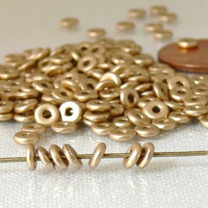 May include: A collection of small, metallic gold-colored donut-shaped beads. Some beads are strung on a thin wire, while others are scattered in a pile. A copper coin is visible in the background.