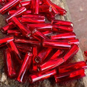May include: A pile of vibrant red, cylindrical glass beads. Each bead has a slightly irregular shape and a glossy finish, reflecting light. The beads are scattered, creating a textured, eye-catching display. These beads are ideal for jewelry making and crafting.
