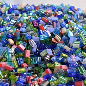 May include: A pile of colorful glass beads in various shapes and sizes. The beads are primarily rectangular and cylindrical, with a mix of colors including blue, green, pink, and clear. Some beads have a metallic sheen.