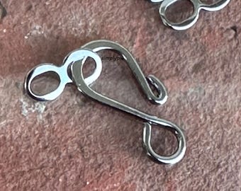 May include: Silver metal clasp with a loop and a hook. The clasp is designed for attaching two pieces of fabric or material together.