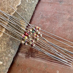 Puede incluir: Una colecci&oacute;n de resistencias axiales con cuerpos cil&iacute;ndricos de color beige y bandas de m&uacute;ltiples colores. Cada componente electr&oacute;nico presenta cables met&aacute;licos largos, delgados y rectos que se extienden desde ambos extremos del cuerpo.