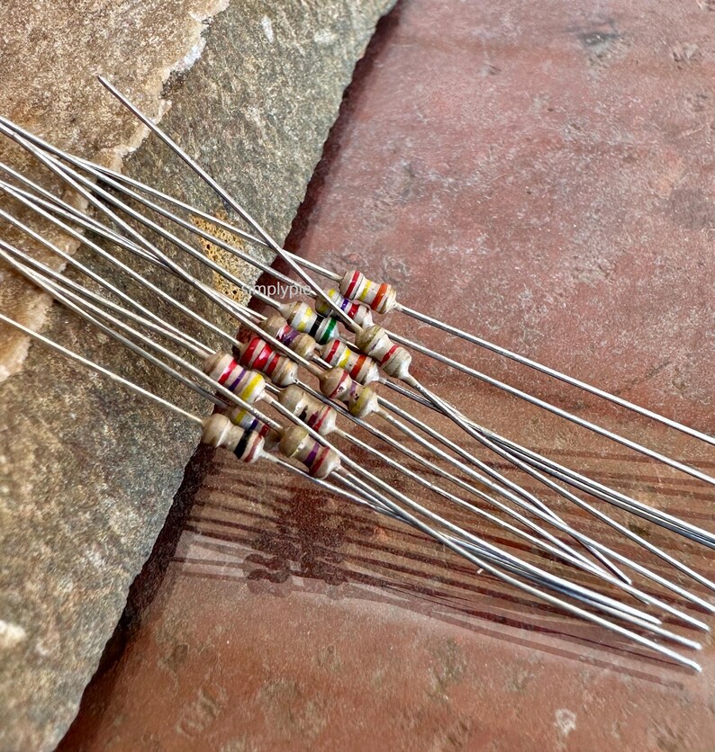 Puede incluir: Una colecci&oacute;n de resistencias axiales con cuerpos cil&iacute;ndricos de color beige y bandas de m&uacute;ltiples colores. Cada componente electr&oacute;nico presenta cables met&aacute;licos largos, delgados y rectos que se extienden desde ambos extremos del cuerpo.